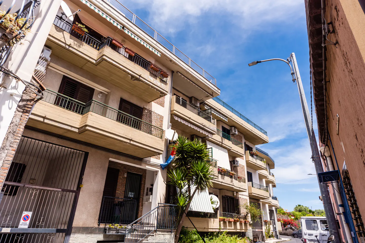 A low-angle shot of a multi-story apartment building with balconies, brown shutters, and a metal gate, under a blue sky.