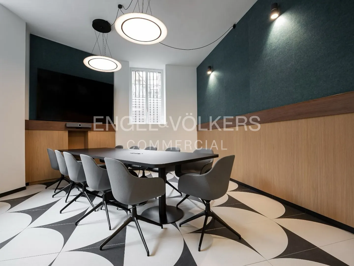 Conference room with a black table, gray chairs, and a large TV screen. The floor has a black and white geometric pattern.