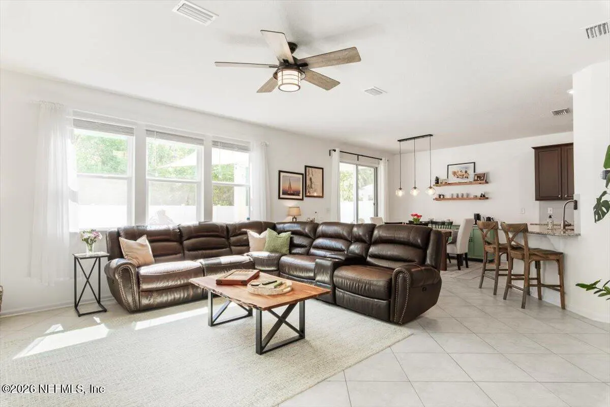 Bright, open-concept living space with a brown leather sectional sofa, wood coffee table, and a ceiling fan. Dining area and kitchen visible in the background.