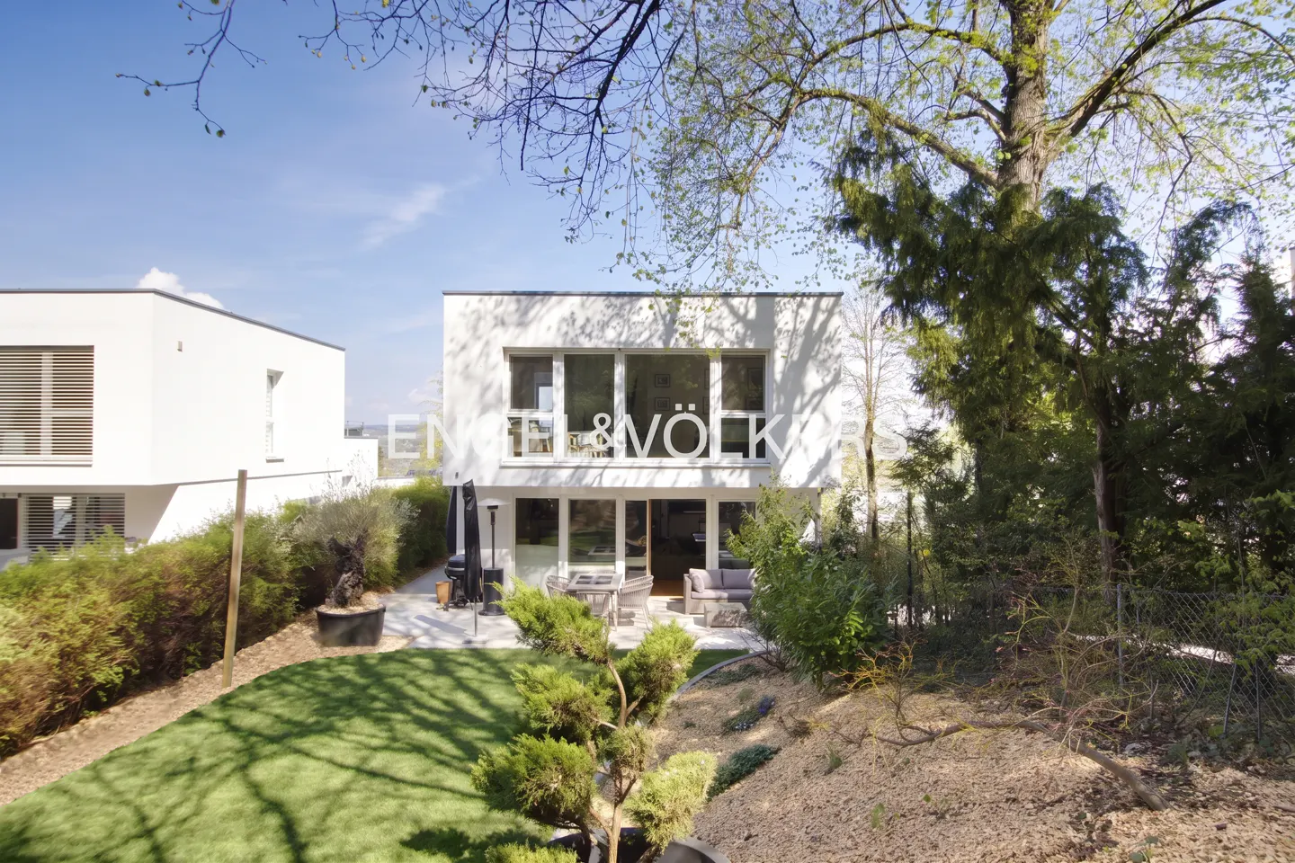 Modern white house with large windows and "Engel & Völkers" logo, surrounded by green lawn, trees, and blue sky.
