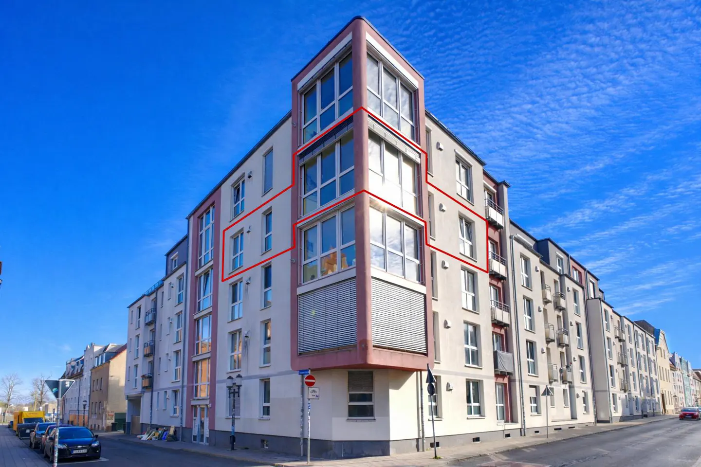 Exterior view of a modern apartment building with large windows and a pink corner tower under a bright blue sky.
