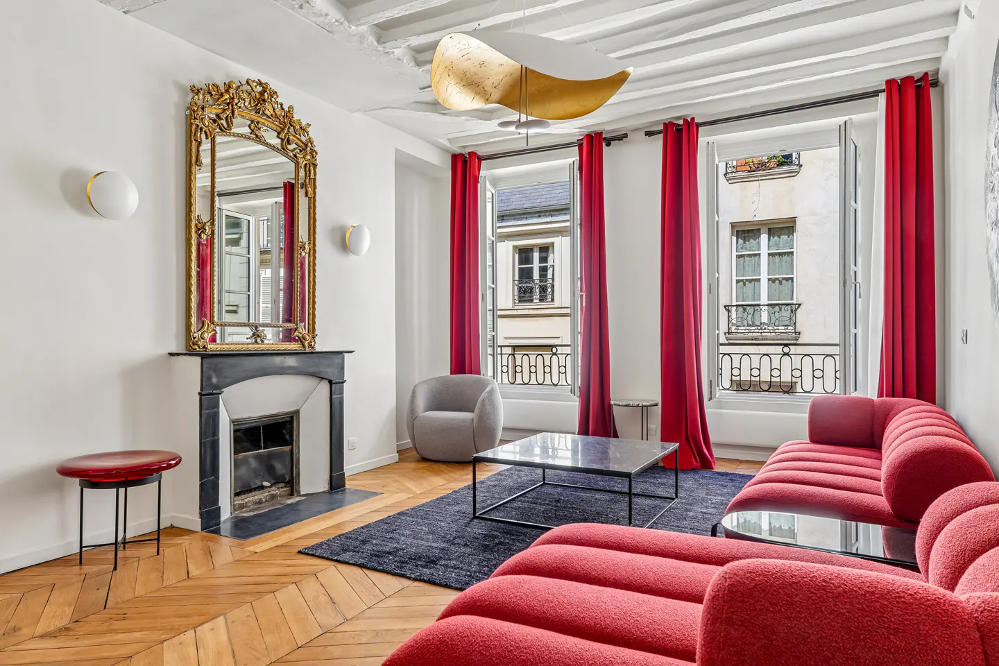 Bright living room with red sofa, black marble fireplace with gold mirror, and open windows with red curtains.