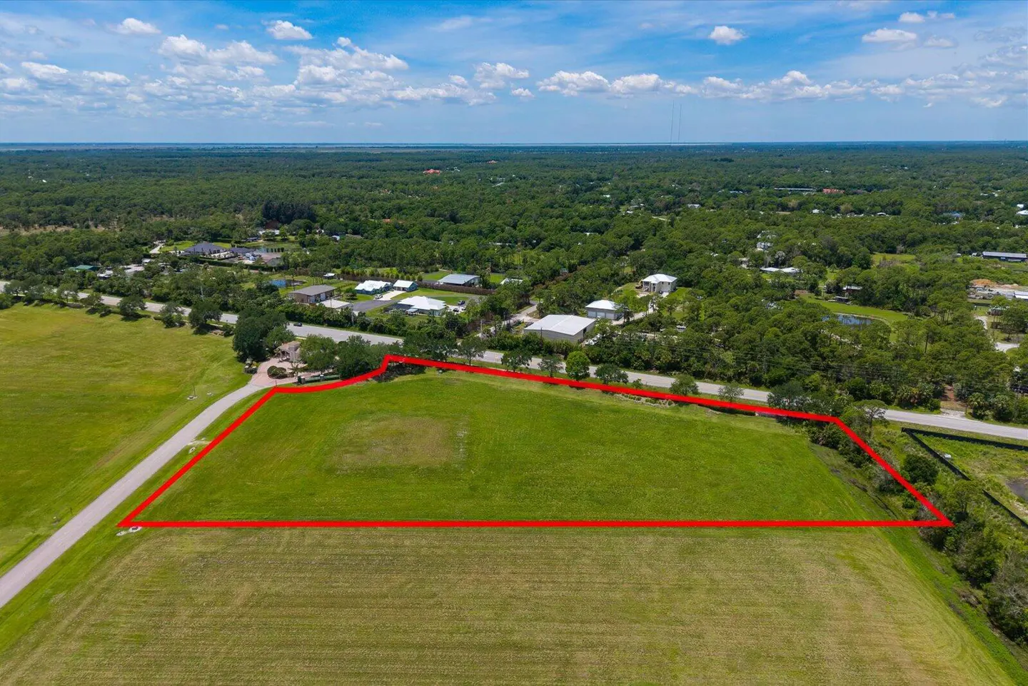 Aerial view of a large, grassy lot outlined in red, with trees, houses, and a blue sky with clouds in the background.
