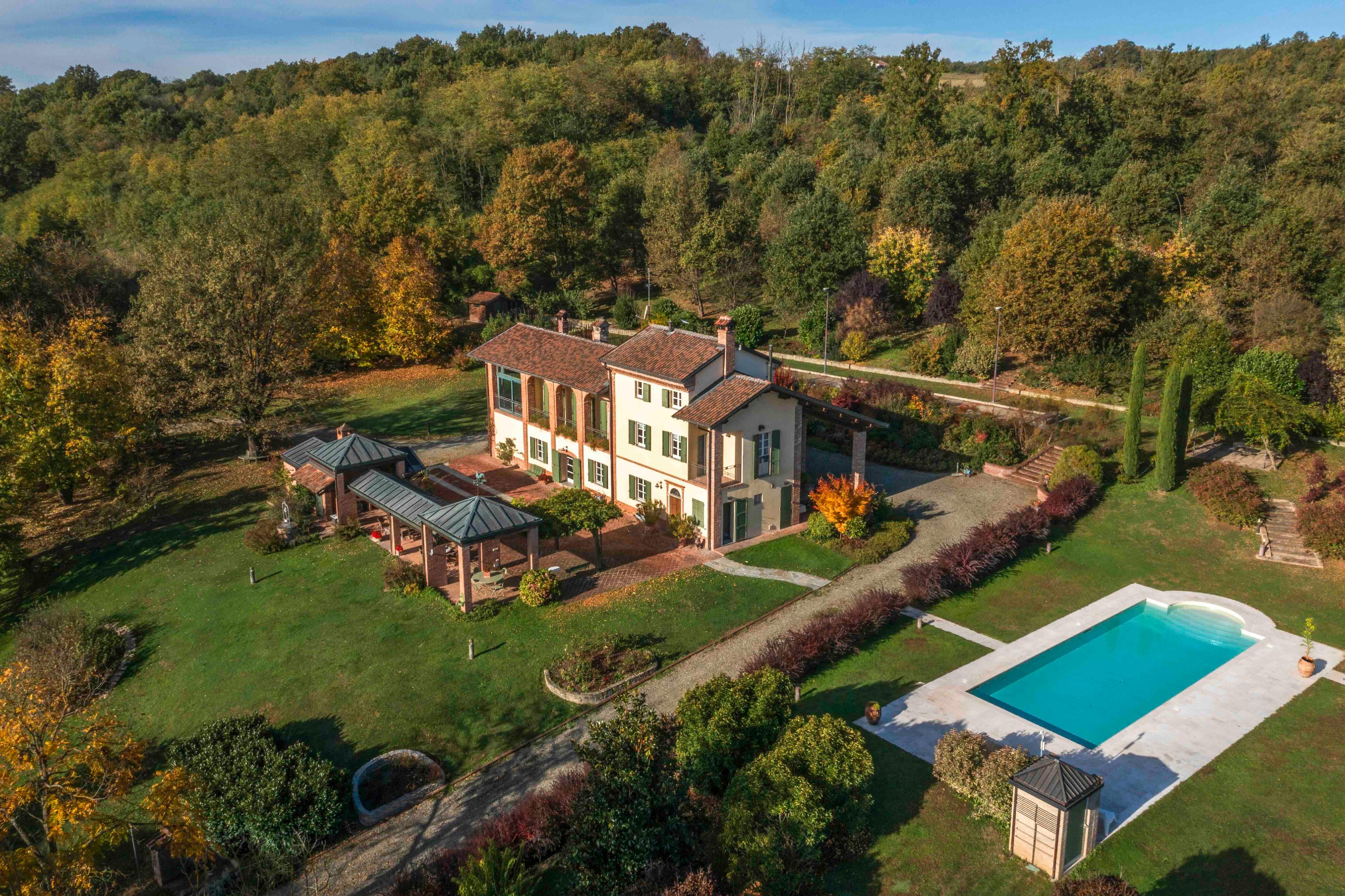 Aerial view of a large, light-yellow house with a red tile roof, green shutters, a pool, and a green lawn surrounded by trees.