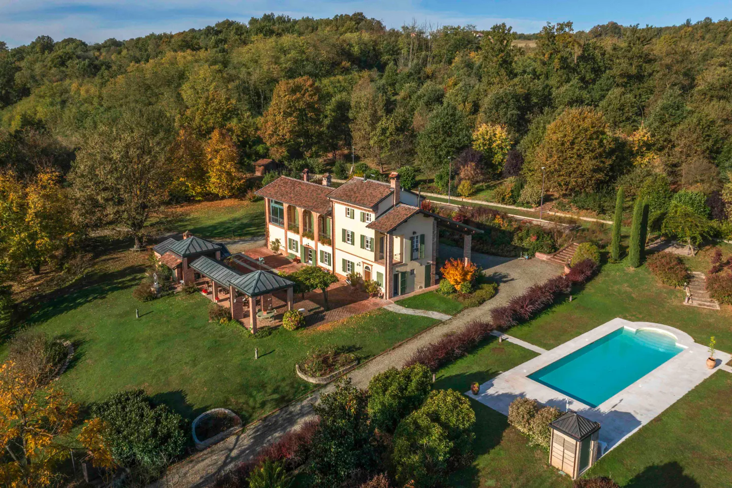 Aerial view of a large, light-yellow house with a red tile roof, green shutters, a pool, and a green lawn surrounded by trees.
