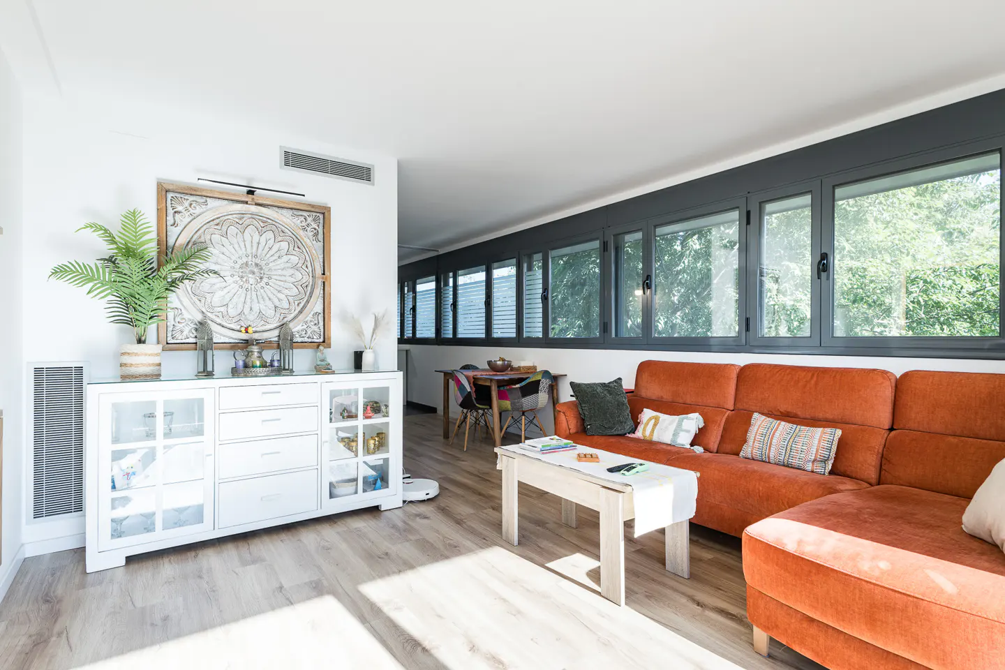 Bright living room with an orange sofa, white cabinet with decor, and a dining table visible in the background.