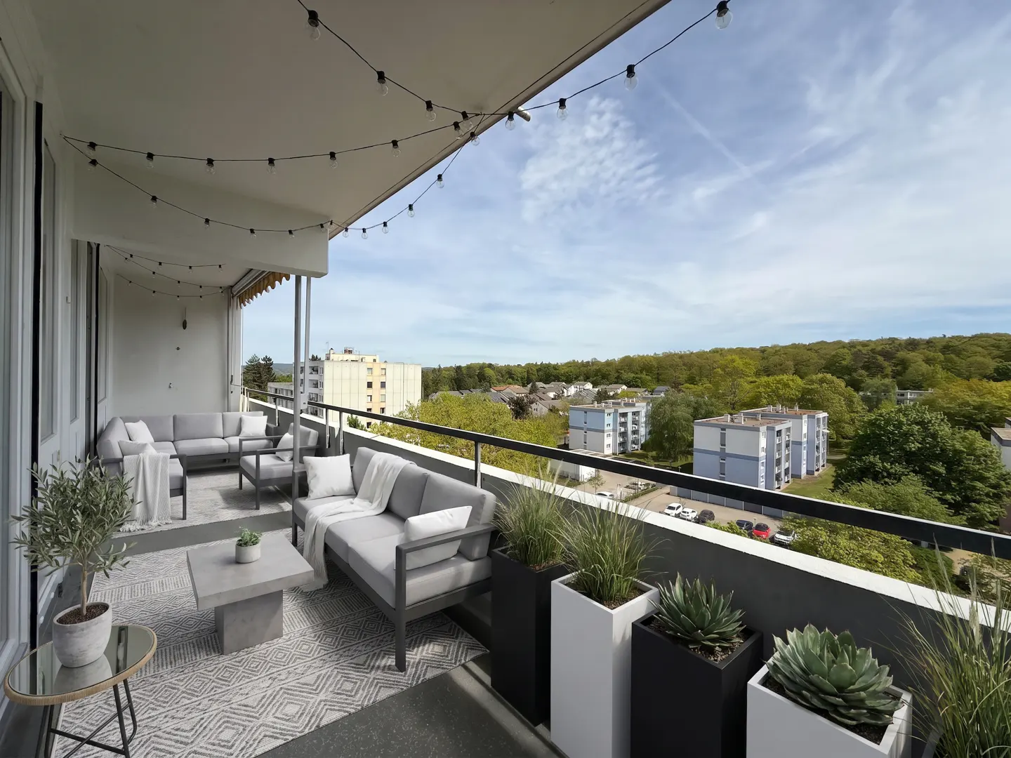 Modern balcony with gray sofas, a concrete coffee table, potted plants, and string lights overlooking a suburban landscape.