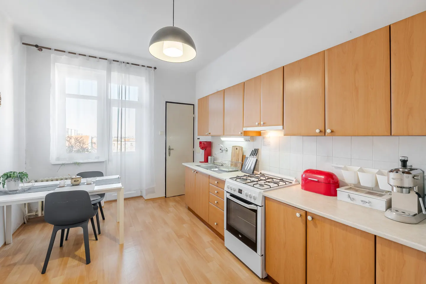 Bright kitchen with light wood cabinets, white appliances, a dining table with two chairs, and a large window with curtains.