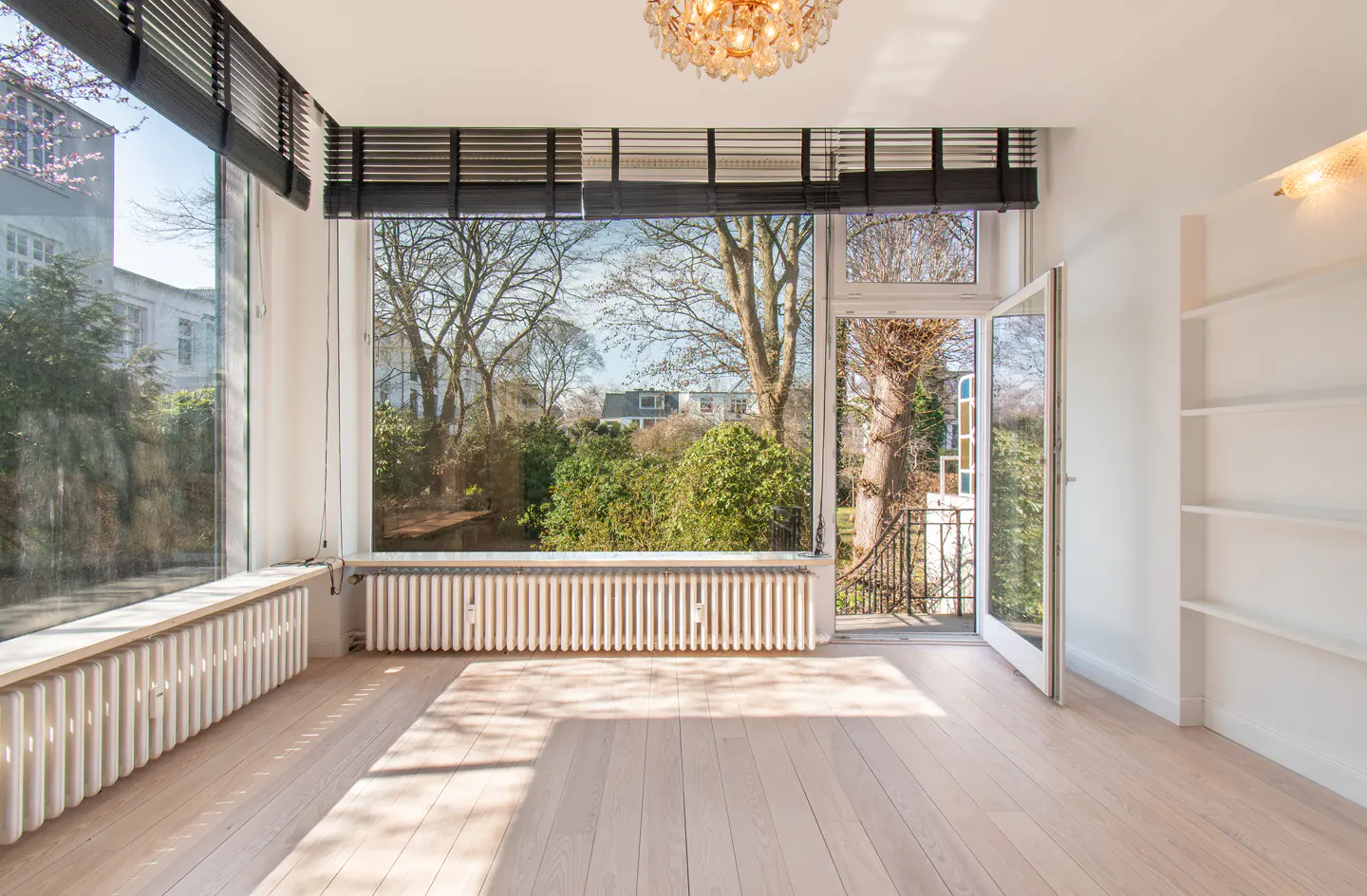 Bright, empty room with light wood floors, white walls, and large windows overlooking a lush green garden. Black blinds are partially closed.