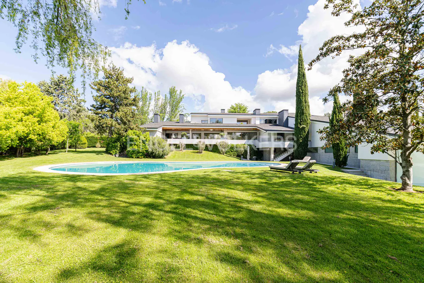 A modern white house with a pool and green lawn on a sunny day. Two lounge chairs sit near the pool.