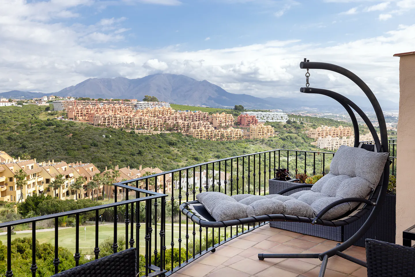 Balcony view with a gray hanging lounge chair. In the background are mountains, green hills, and buildings under a cloudy sky.