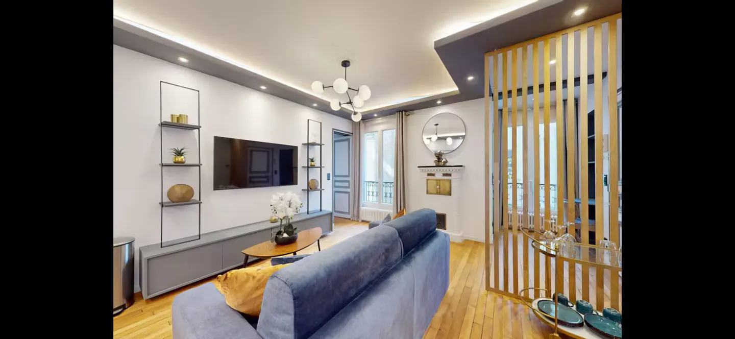 Living room with blue sofa, wood floors, and white walls. A TV hangs above a gray console table with shelves on either side. A gold screen divides the room.