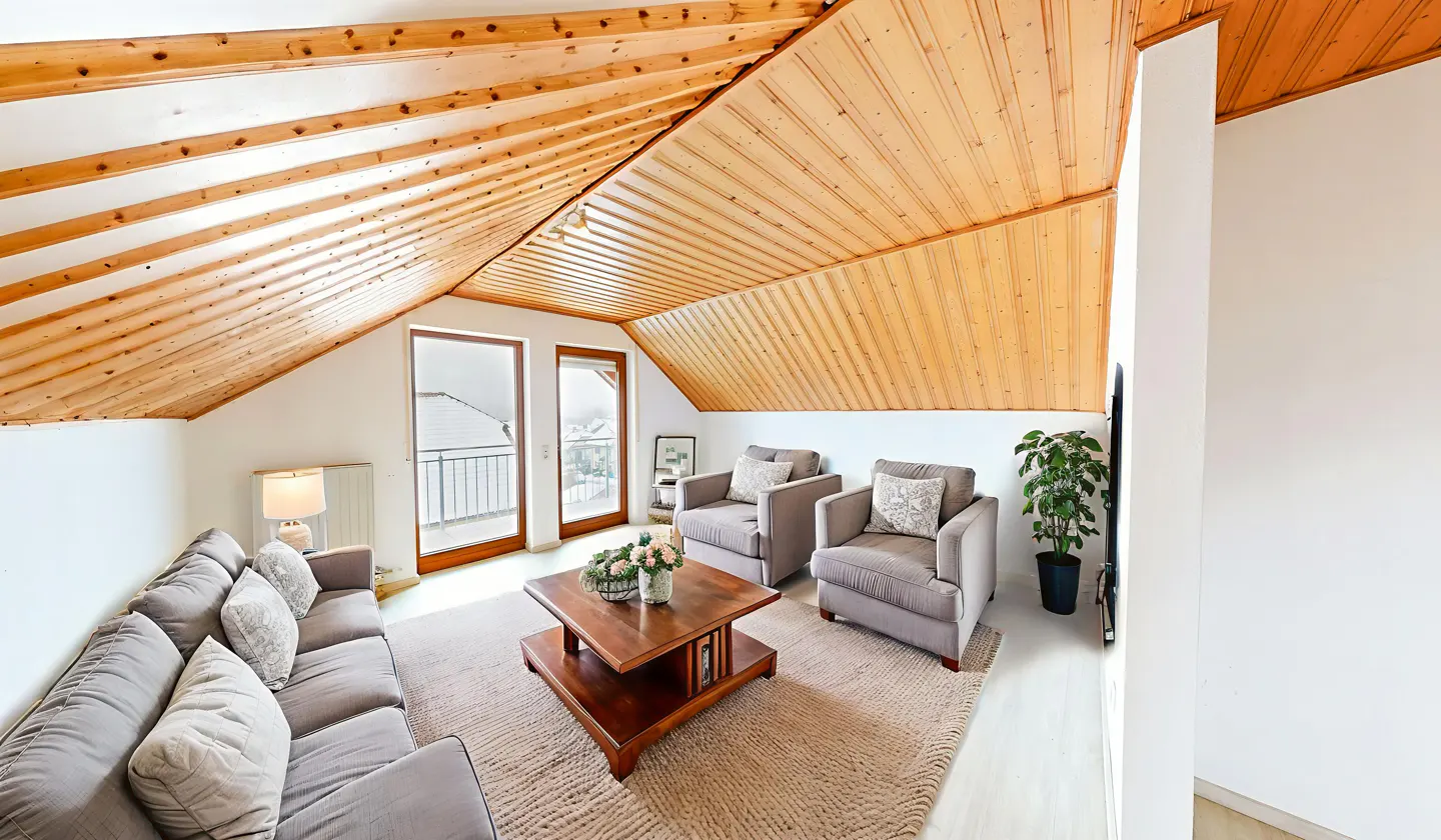 Attic living room with wood-paneled vaulted ceiling, gray sofa, two armchairs, wooden coffee table, and balcony doors.