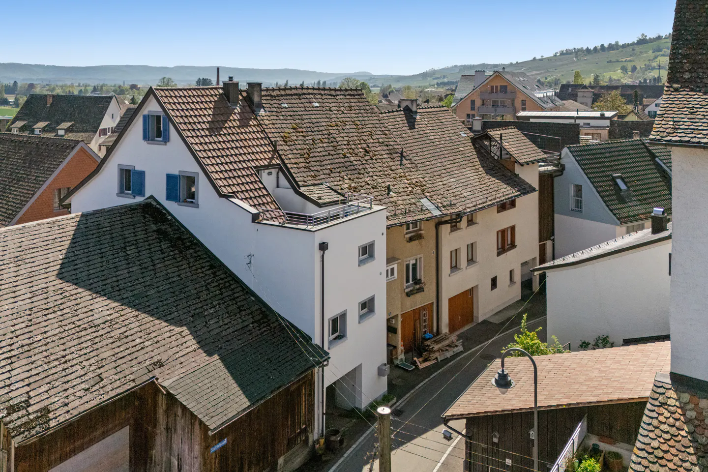 Aerial view of a European town with houses, tile roofs, and a narrow street on a sunny day. Hills are visible in the background.