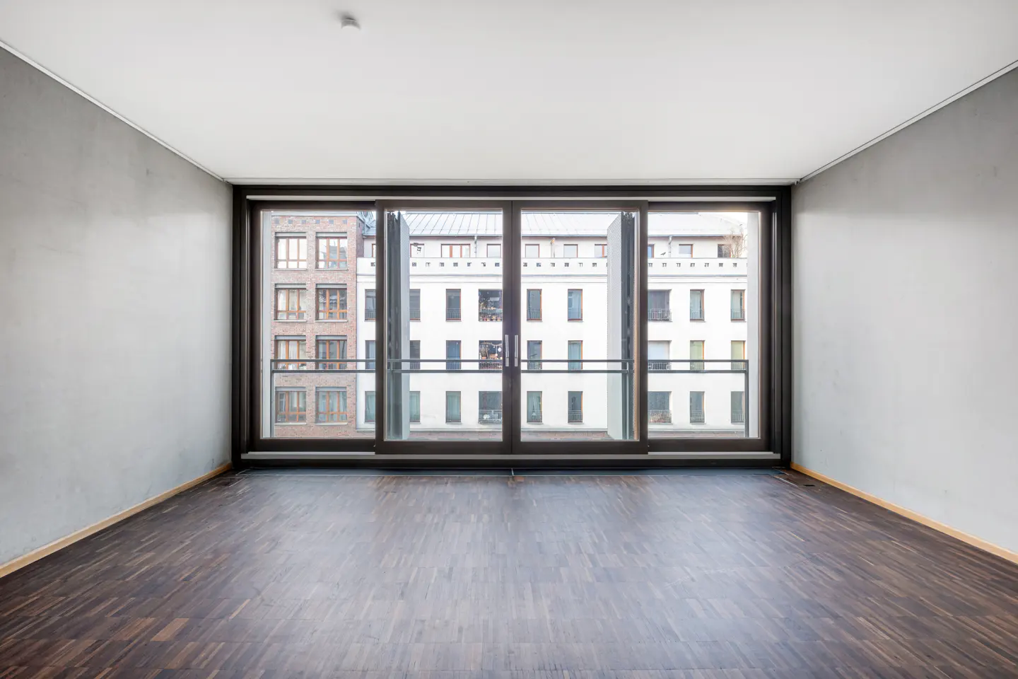 Empty room with gray walls, dark wood floors, and a large window showing buildings outside.