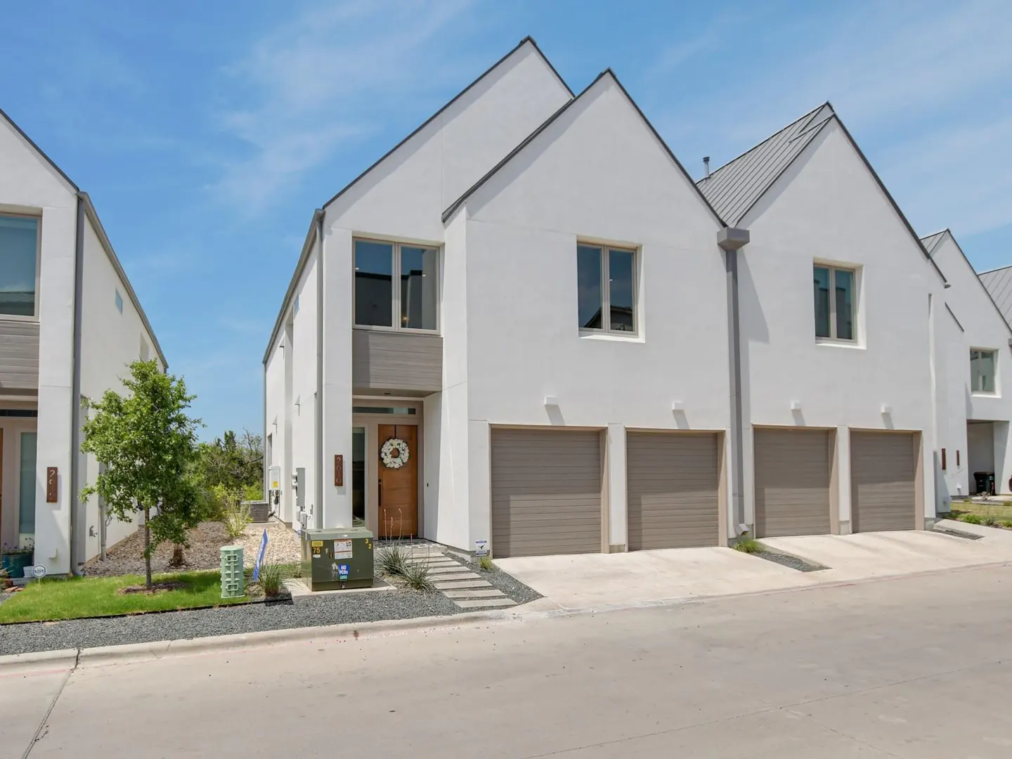 Row of modern white townhouses with gray roofs and garage doors under a blue sky. A wreath hangs on the front door.