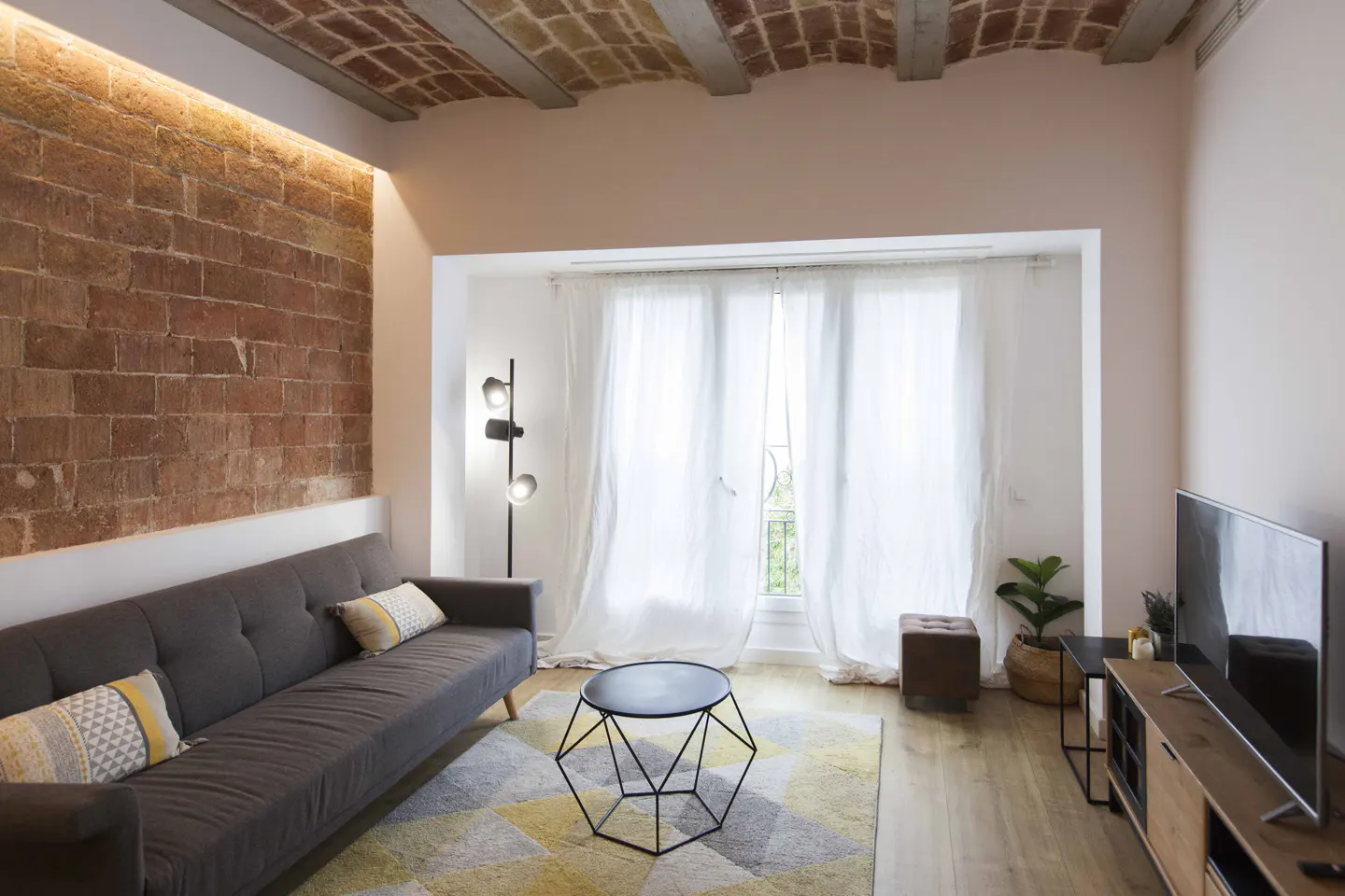 Living room with brick wall, grey sofa, geometric rug, and natural light from window with sheer white curtains.