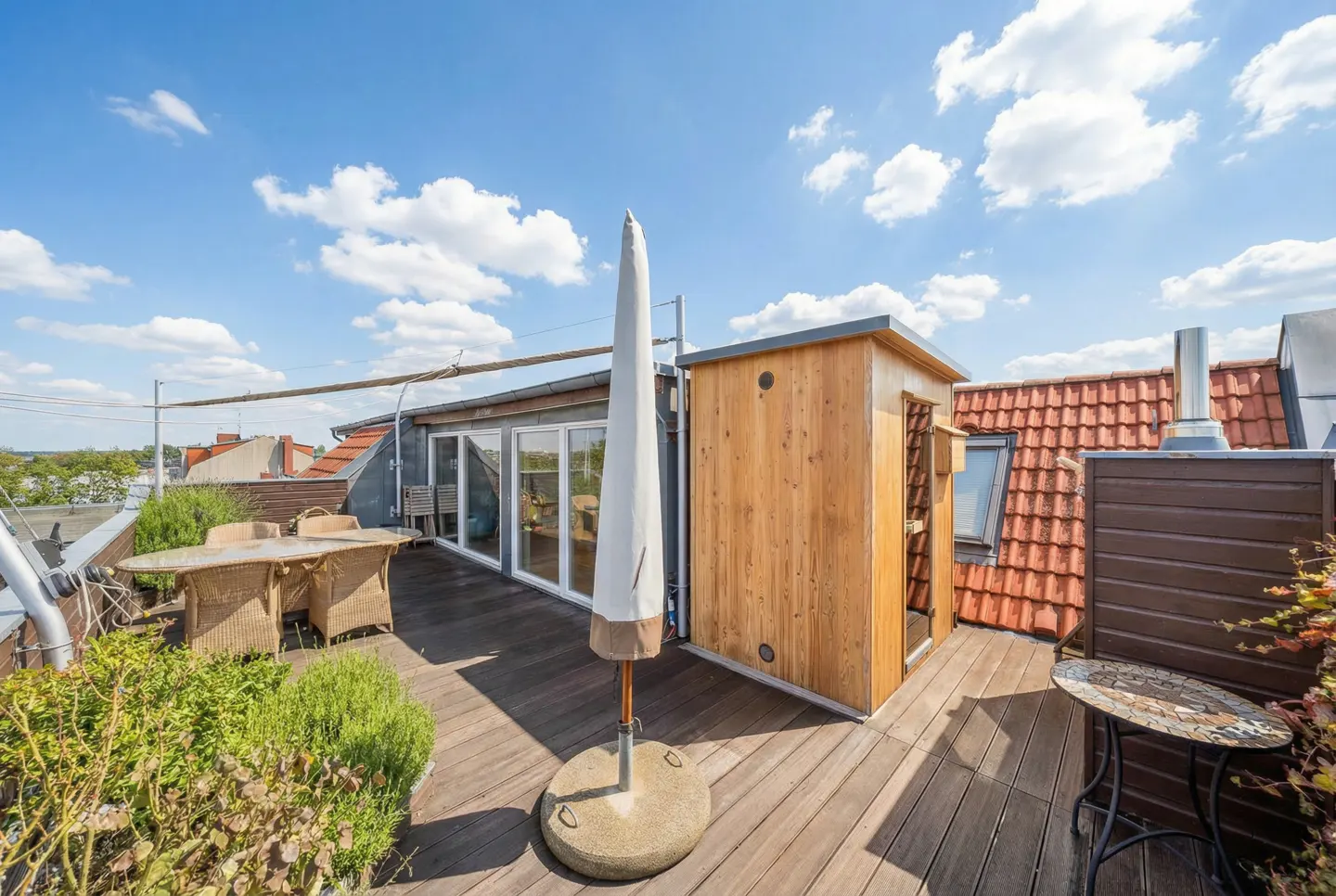 Rooftop deck with wood flooring, table, chairs, closed umbrella, and small wood building under a blue sky with clouds.