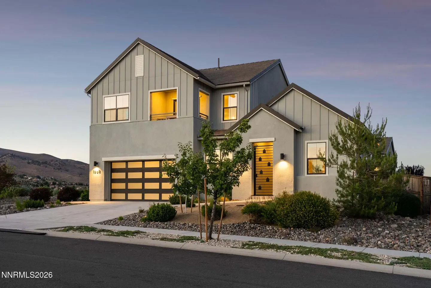 Modern two-story gray home with a multi-car garage, illuminated windows, and desert landscaping at twilight.