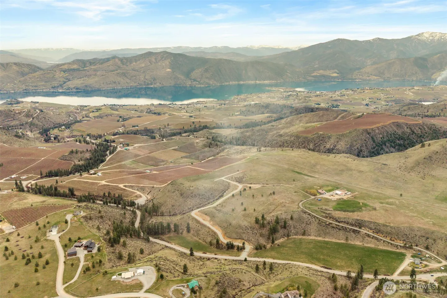 Aerial view of a valley with vineyards, hills, and a lake in the background under a blue sky.