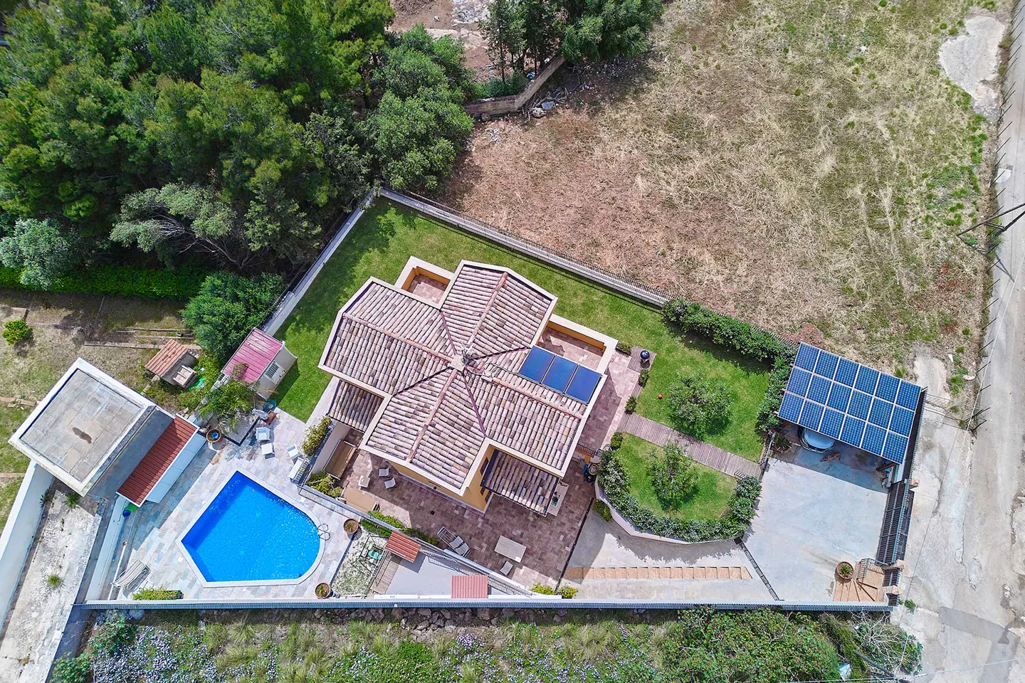 Aerial view of a tan house with a red tile roof, blue pool, solar panels, green lawn, and trees.