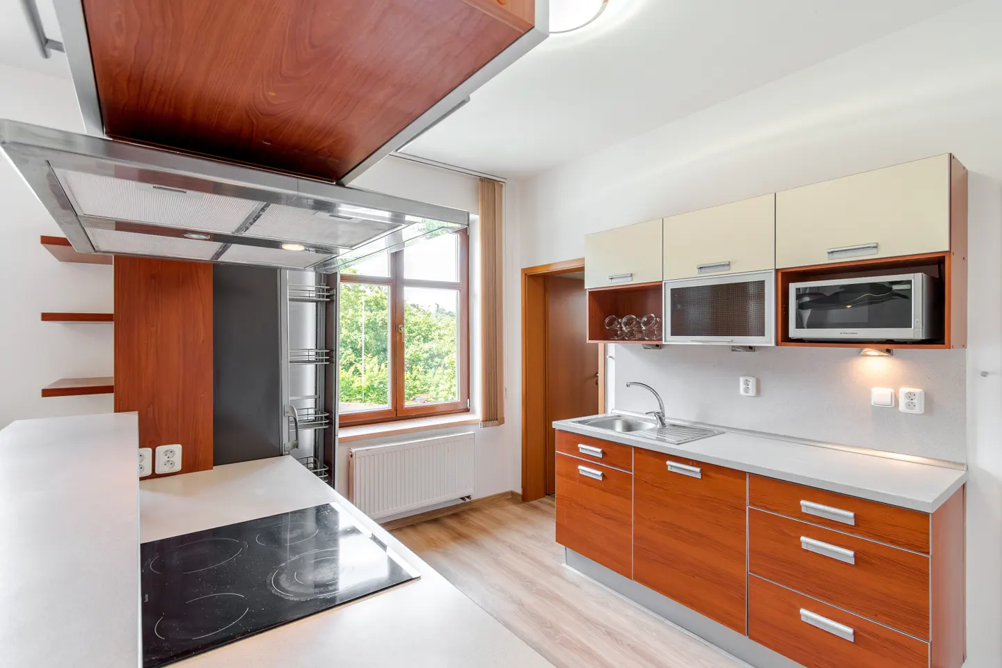 Modern kitchen with wood-tone cabinets, white countertops, a black induction cooktop, and a window overlooking greenery.