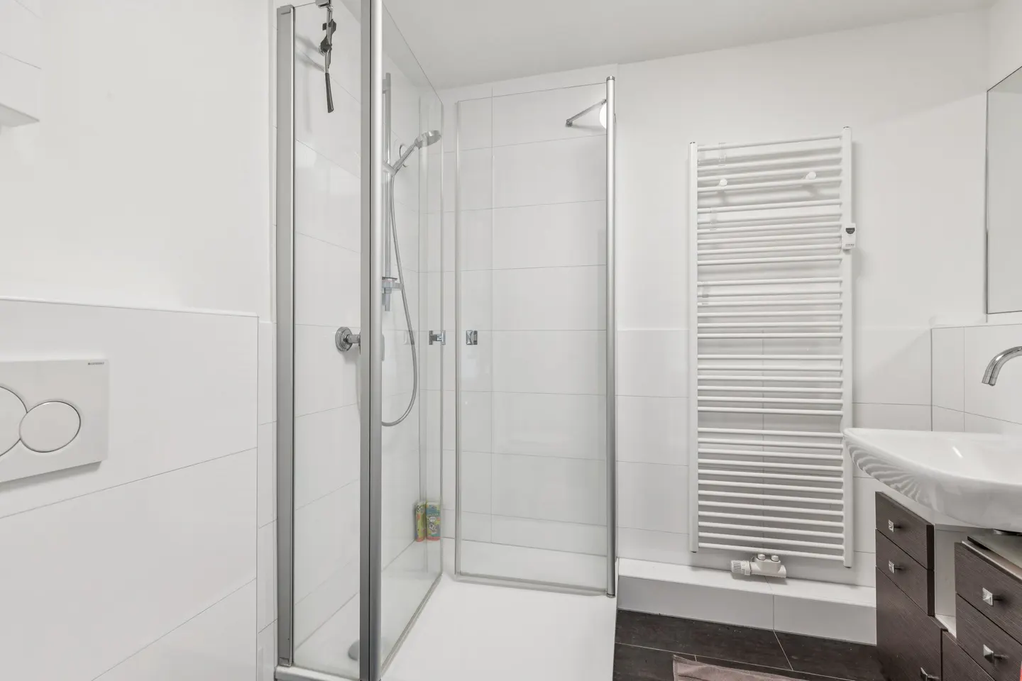 Bright bathroom with a glass shower stall, white tiled walls, and a white towel warmer. A dark vanity with a white sink is visible.