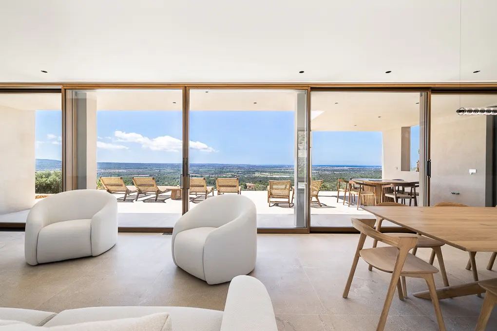 Bright, modern living room with white chairs, a wooden table, and sliding glass doors to a patio with lounge chairs and a scenic view.