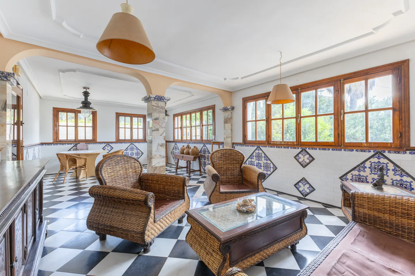 Bright sunroom with wicker furniture, black and white checkered floor, and blue and white tiled walls. Windows look out to green trees.