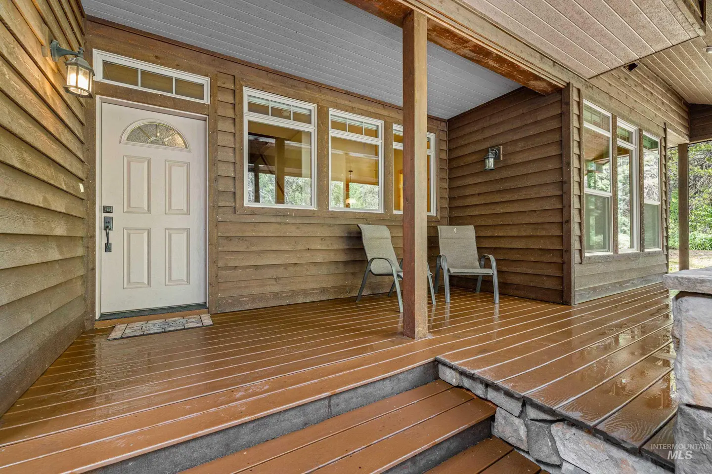Exterior view of a brown wood-sided house with a covered porch, white door, and two chairs.