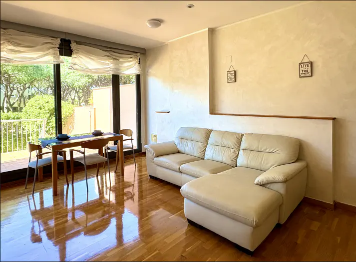 Living room with hardwood floors, a beige leather sofa, and a dining table with chairs near a large window.