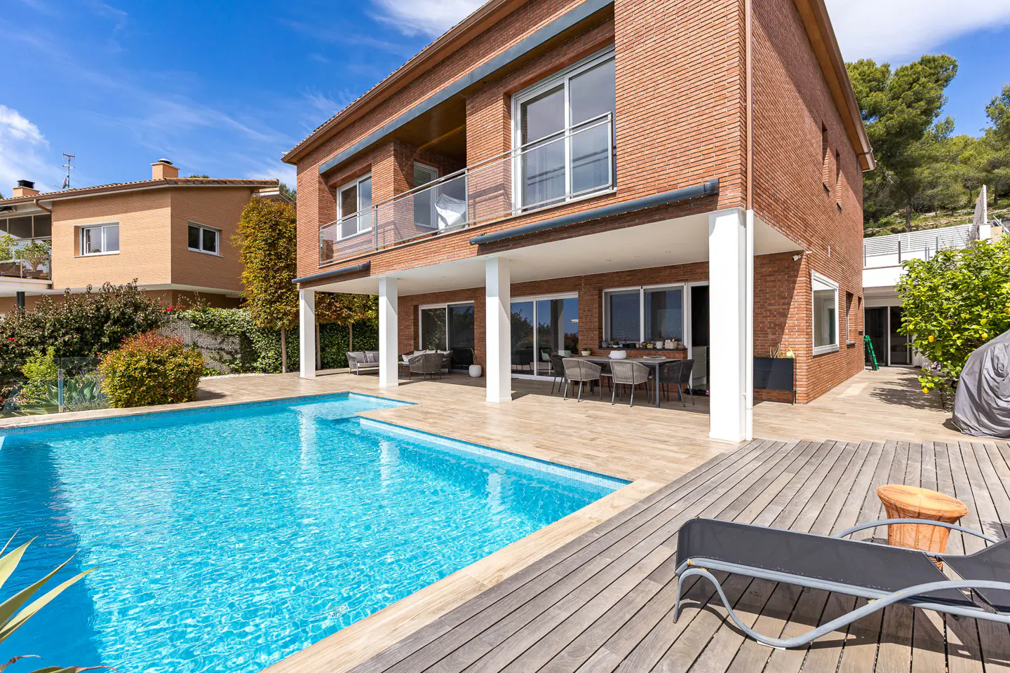 Brick house with a blue pool and wooden deck. Patio furniture is visible under a covered porch.