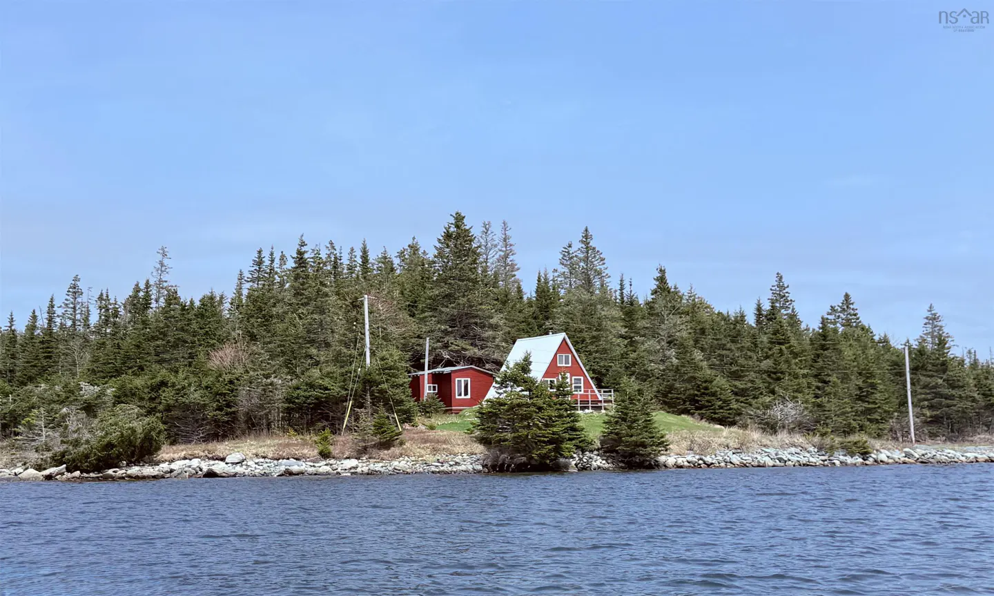 Red A-frame house on a small island covered in evergreen trees, viewed from the water under a clear blue sky.