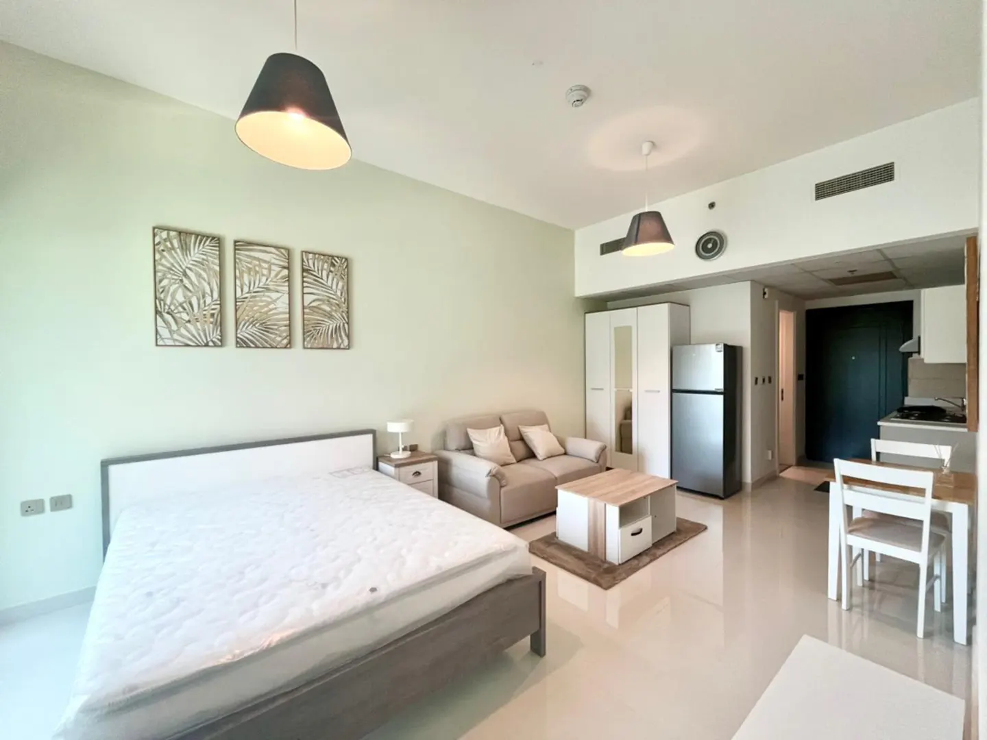 Studio apartment with a bed, sofa, table, and kitchen area. The walls are light green, and there are three leaf-themed pictures on the wall.