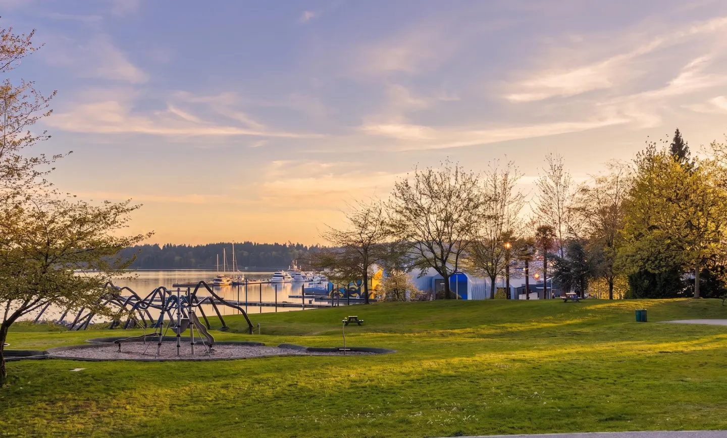 Park view with green grass, trees, and a playground. Boats are docked in the water in the background under a pastel sky.