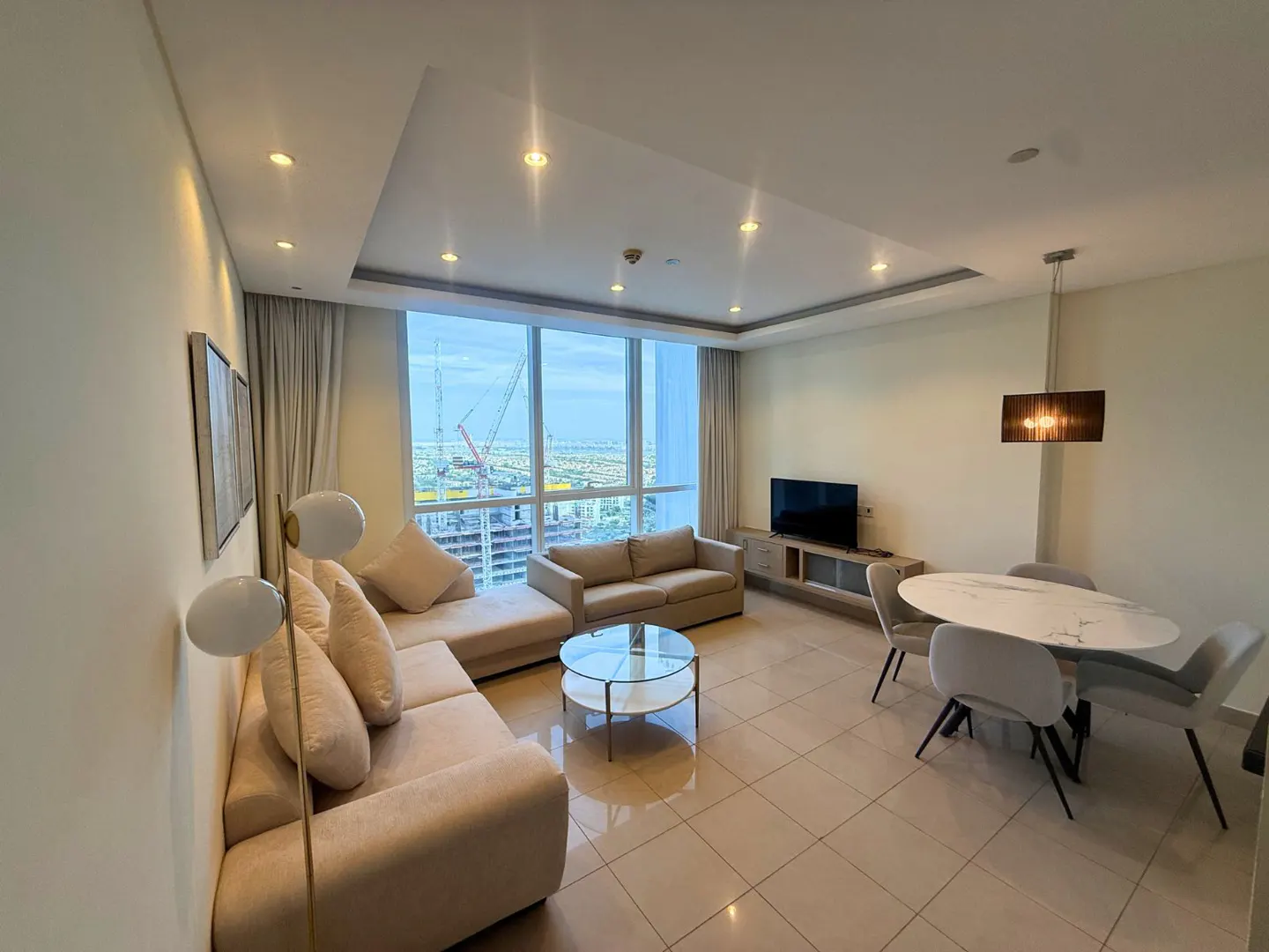 Bright living room with beige sofas, a glass coffee table, and a dining area with a marble table. Large windows offer a city view with a construction crane.