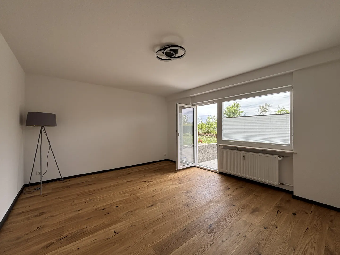 Bright, empty room with wood floors, white walls, and a modern light fixture. A floor lamp stands in the corner, and a door opens to a balcony.