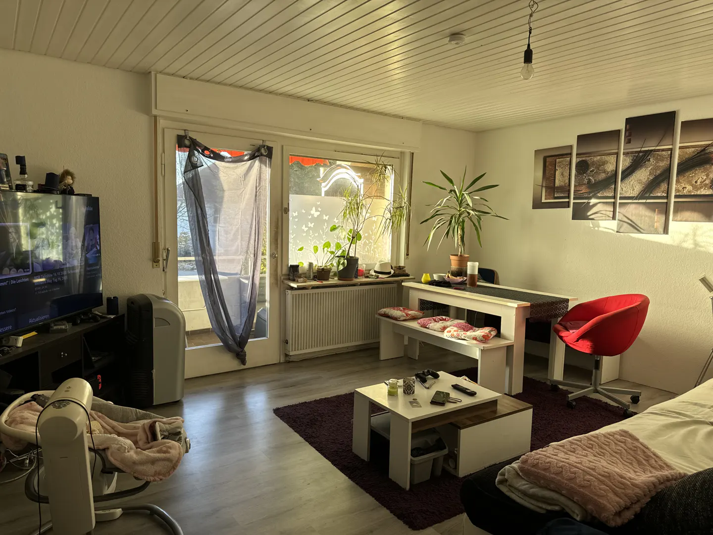 A bright living room with a TV, dining table, red chair, and a baby swing. Sunlight streams through the windows.