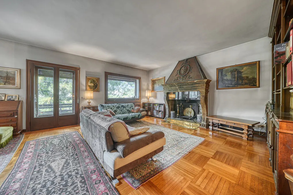 Living room with parquet floors, sofas, and a large ornate fireplace. A rug covers part of the floor. Bookshelves and paintings decorate the walls.