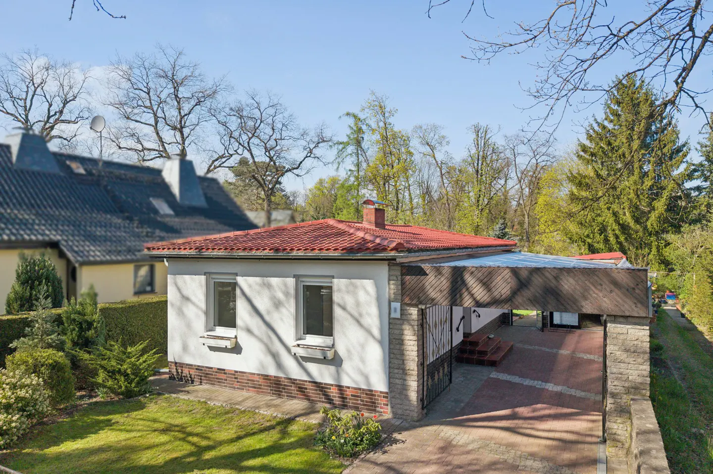 A single-story white house with a red tile roof and a carport, surrounded by green grass and trees.