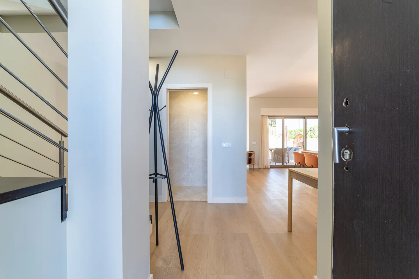 View from an open dark door into a bright home interior with light wood floors, a coat rack, and a view of a patio.