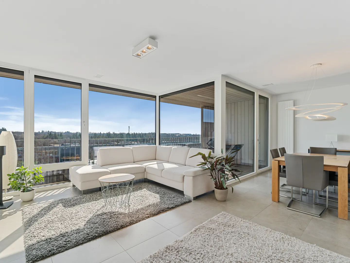 Bright, modern living room with a white sectional sofa, large windows, and a dining area with gray chairs.