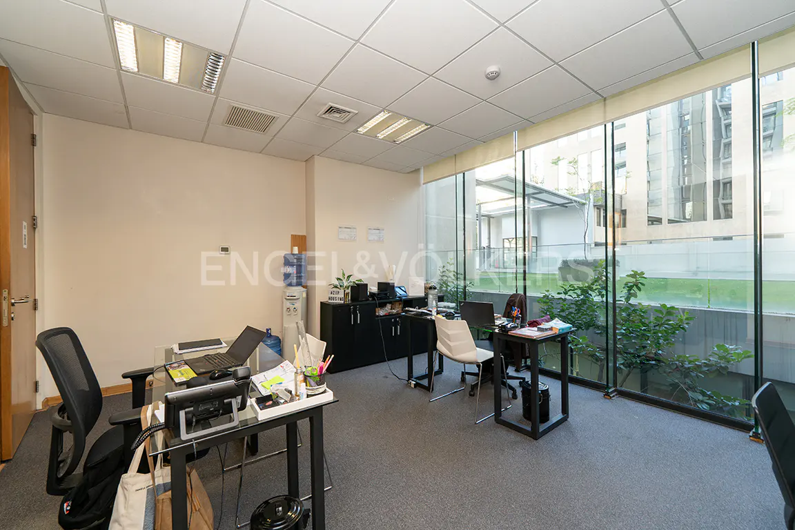 Bright office space with desks, chairs, and large windows overlooking greenery and buildings. Neutral walls and gray carpet. Engel & Völkers logo visible.