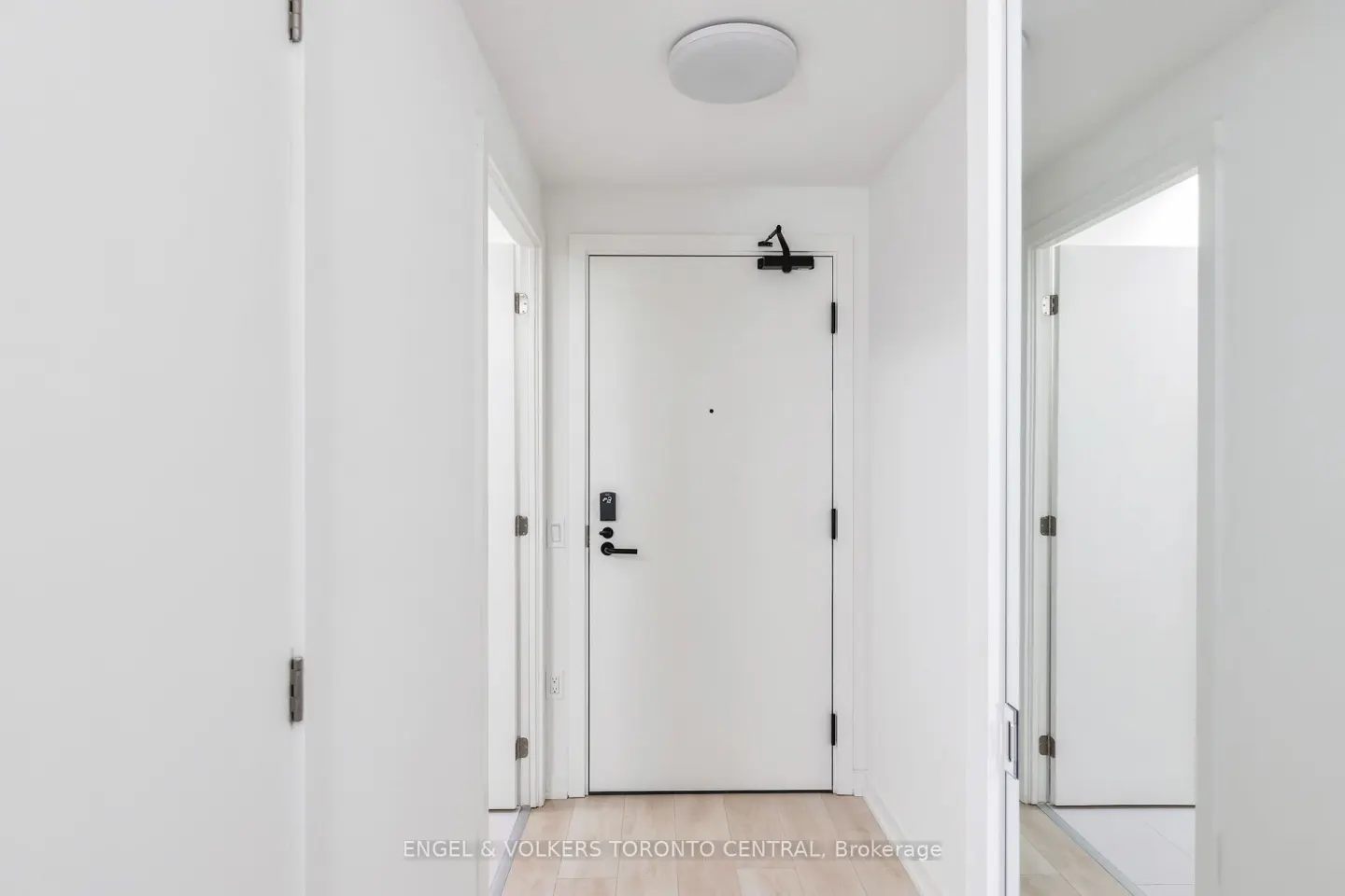 Bright, narrow hallway with white walls, doors, and light wood floors. A white front door with black hardware is centered.