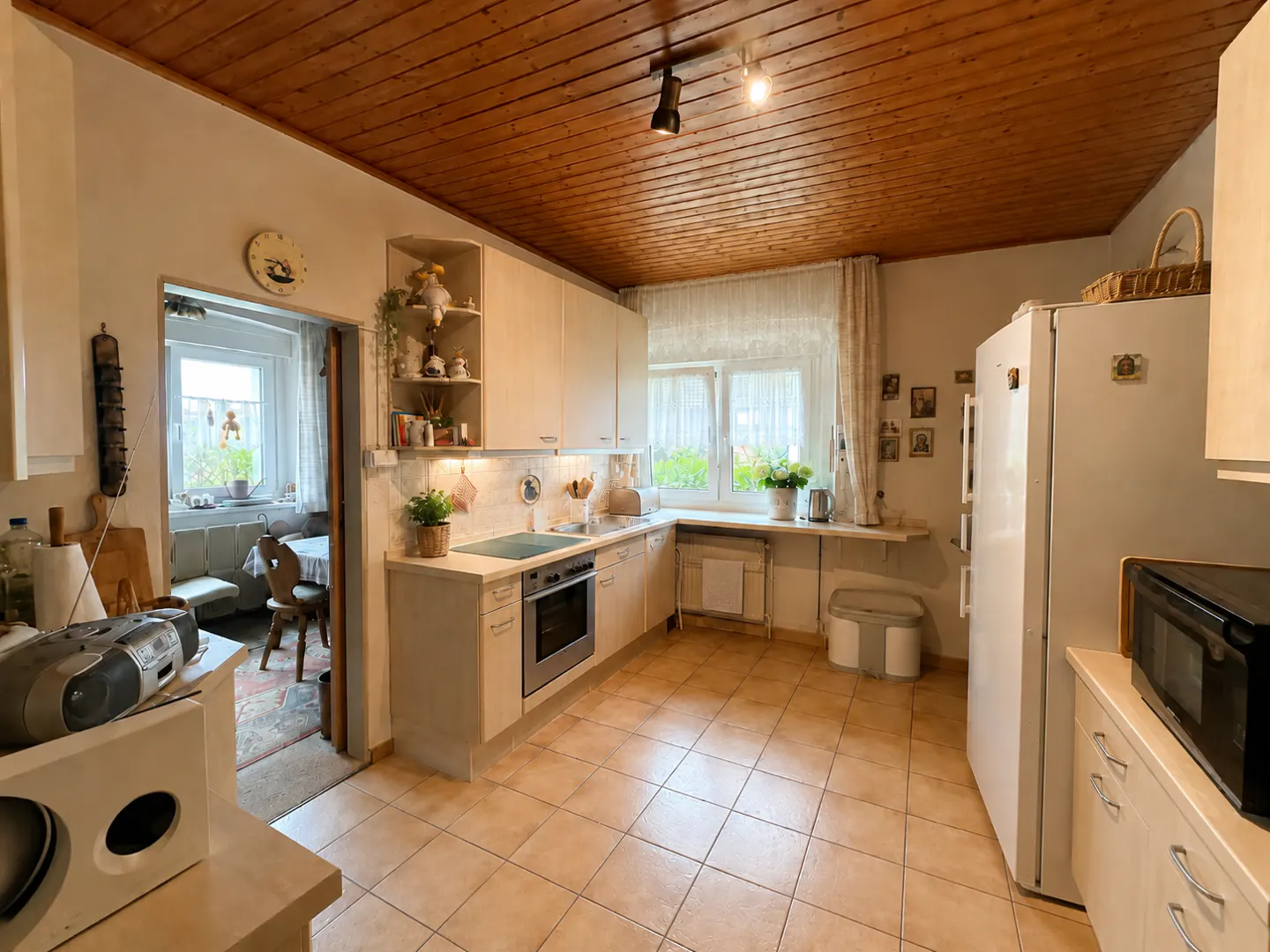 A bright kitchen with beige cabinets, a wooden ceiling, and tile floors. A window with sheer curtains lets in natural light. A dining area is visible through an open doorway.