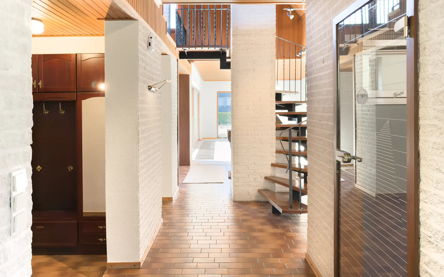 Hallway with brick floors, white brick walls, and wood ceiling. A spiral staircase and a glass door are visible. A dark wood cabinet is on the left.