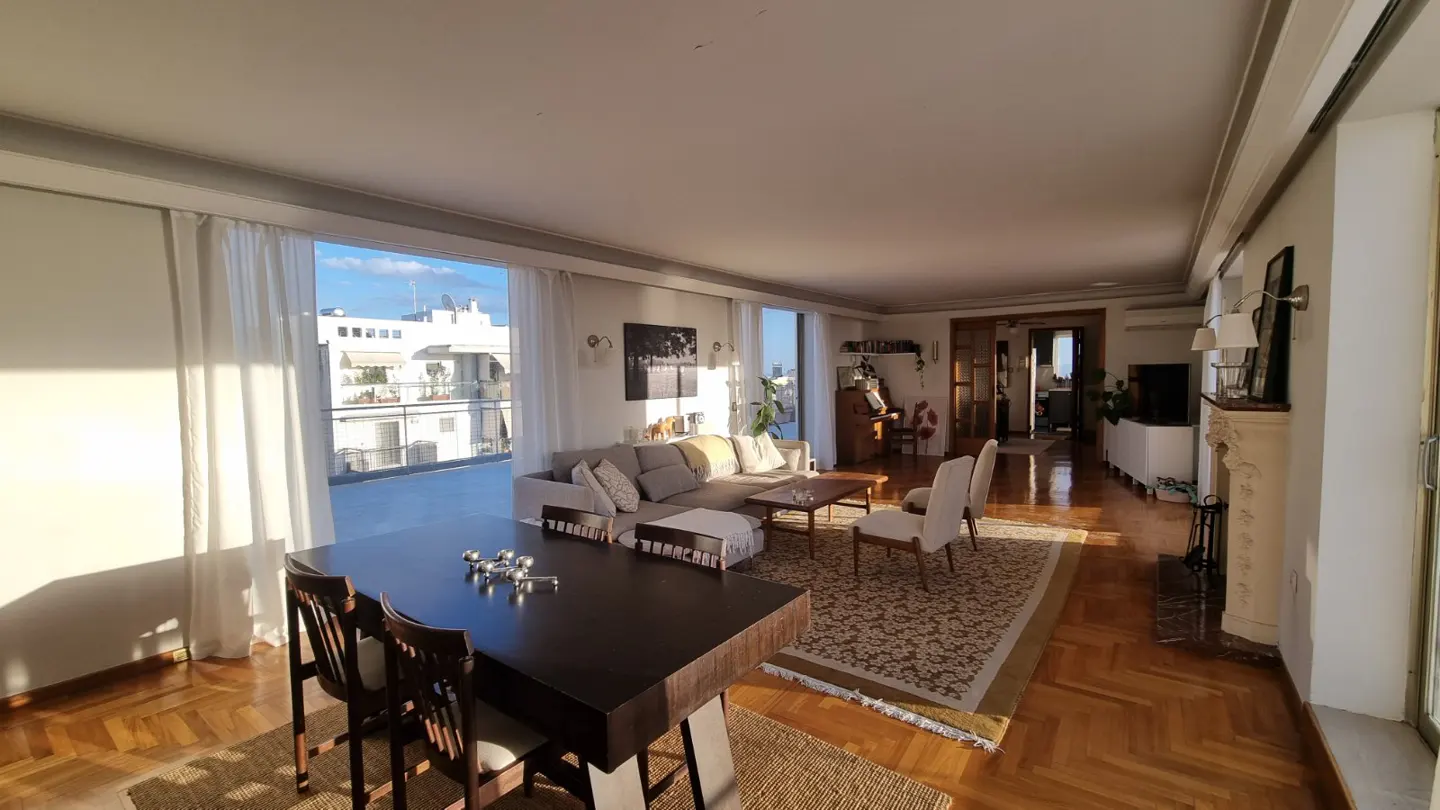 Bright, open-plan living room with a dark wood dining table, beige sofa, and herringbone wood floors. Balcony with city views visible through sheer curtains.