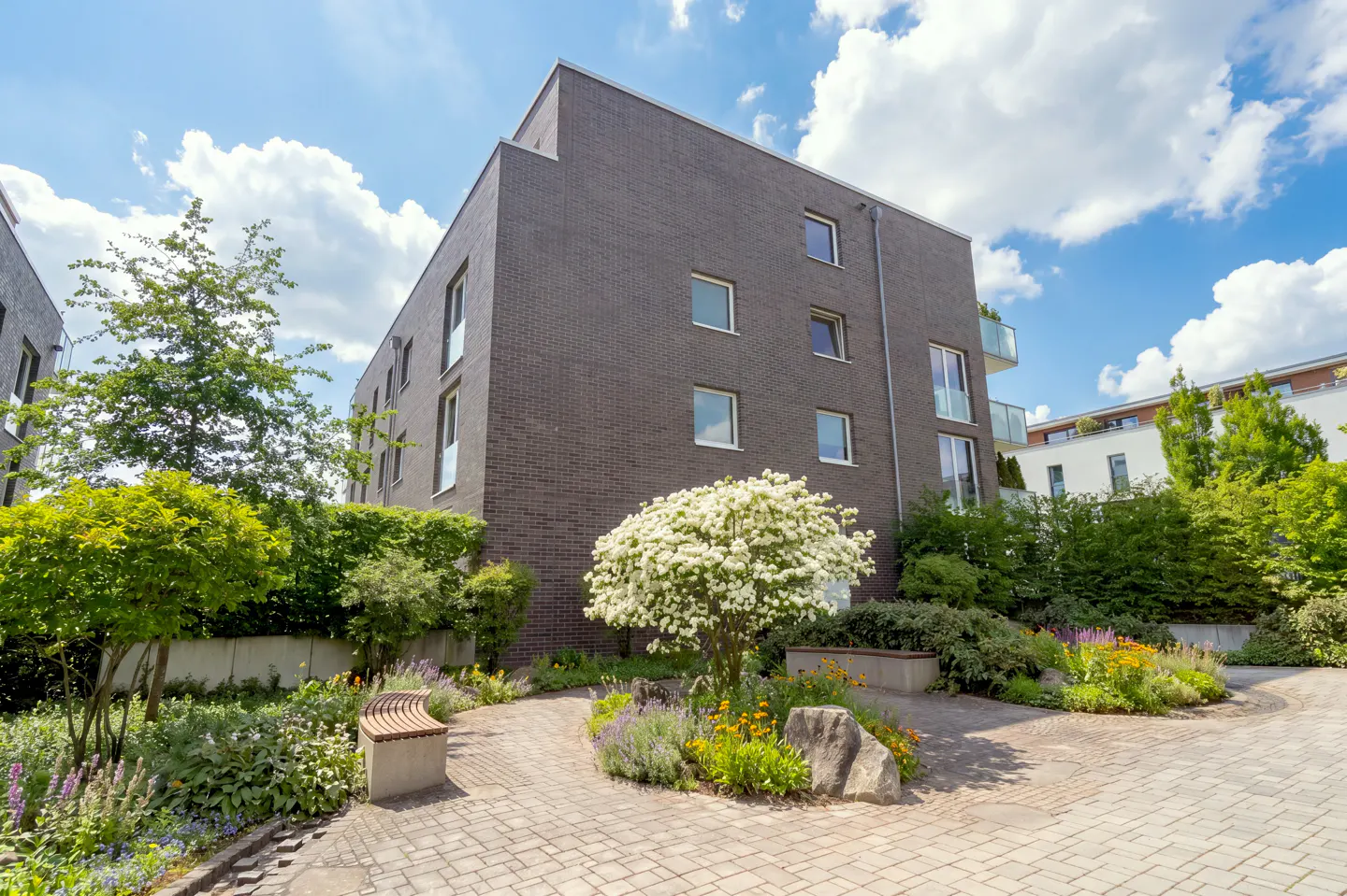 Modern brick apartment building with a landscaped courtyard, featuring a bench, flowers, and a small tree with white blossoms.