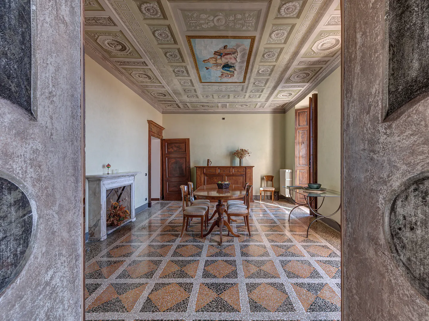 Ornate room with patterned tile floor, wood table and chairs, fireplace, and painted coffered ceiling.