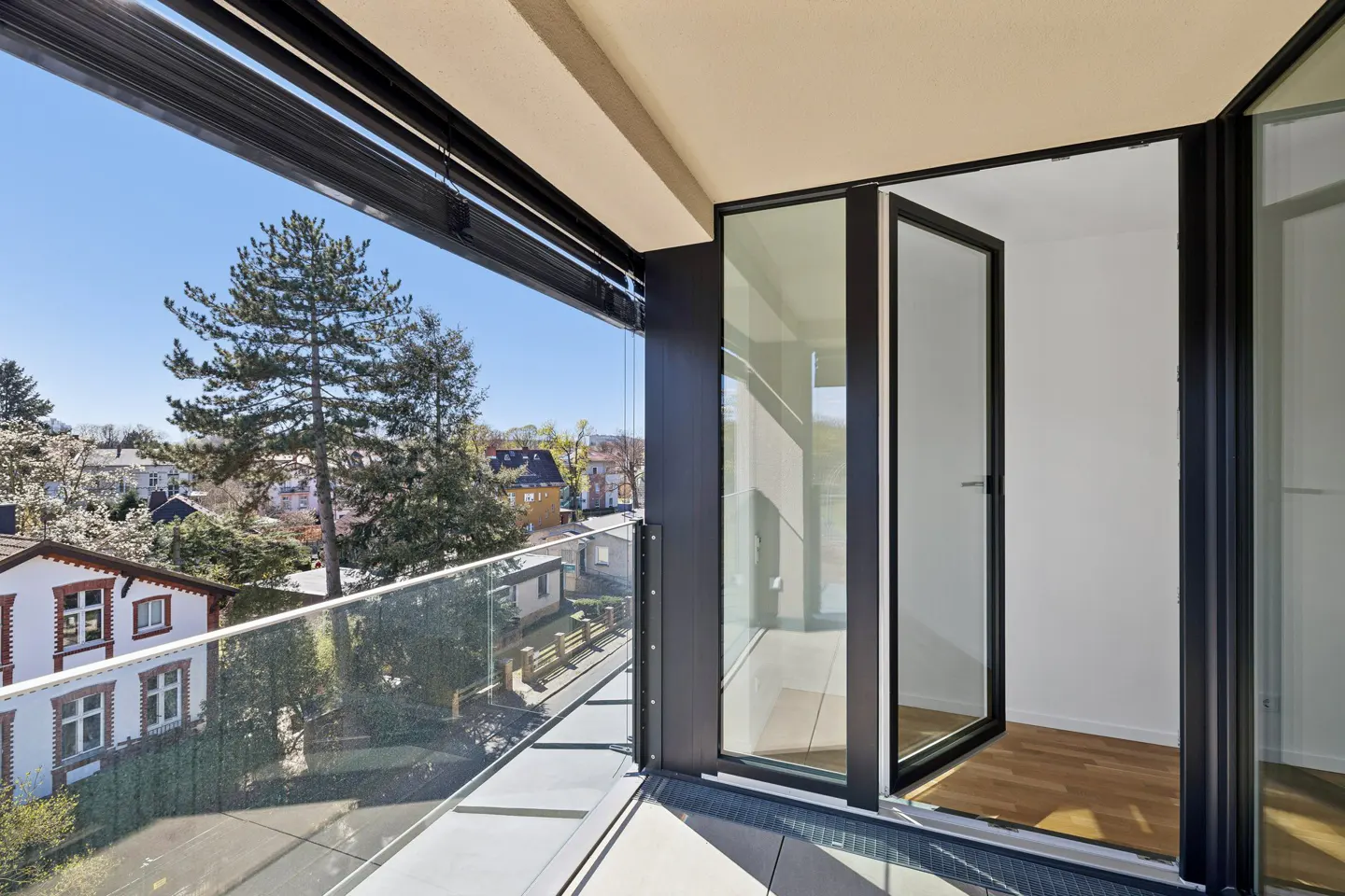Balcony with glass railing and black frame sliding doors open to a room with wood floors, overlooking trees and houses on a sunny day.